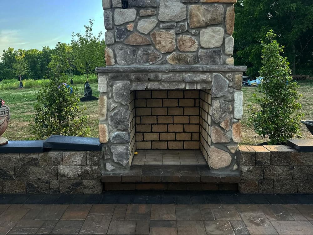 Stone outdoor fireplace with empty hearth, surrounded by landscaping and greenery.