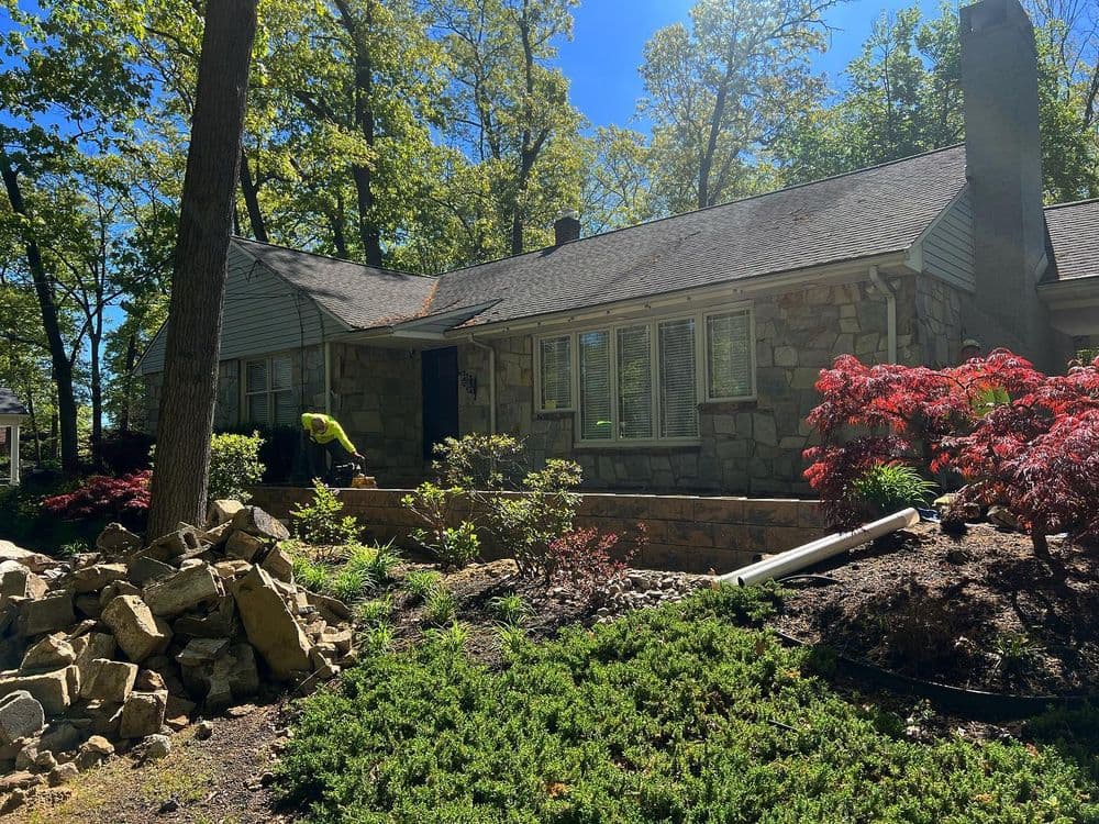 Stone house surrounded by greenery, with a worker gardening in bright sunlight.
