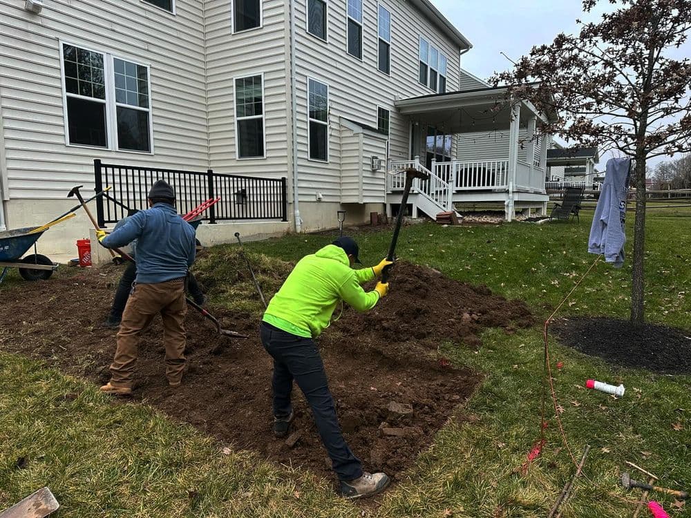 Workers digging a garden plot in a residential backyard near a white house.