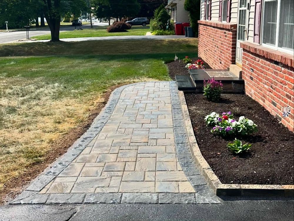 Paved pathway leading to a brick house with flower beds and green lawn.